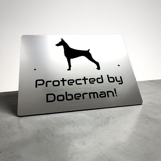 Strieborná tabuľka so siluetou dobermana a anglickým nápisom Protected by Doberman